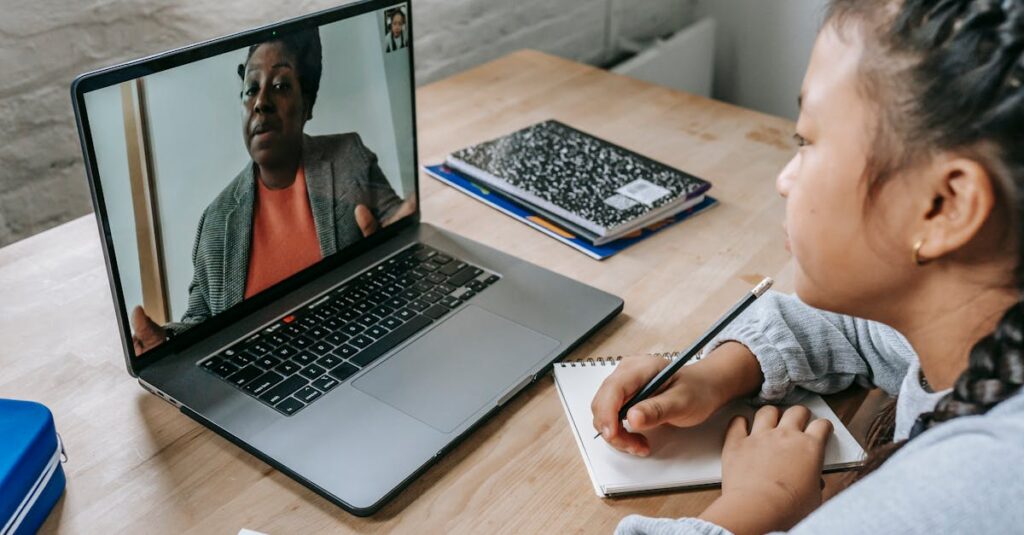 From above of focused Asian girl sitting at table with copybook and pencil and carefully listening to black teacher explaining homework task during videocall on laptop