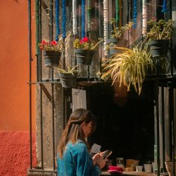 A woman stands by a vividly decorated window in San Miguel de Allende, Mexico, bathed in sunlight.